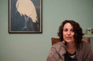 photo of shari, a white woman with short dark curly hair, against a green wall, with a bird painting in the background